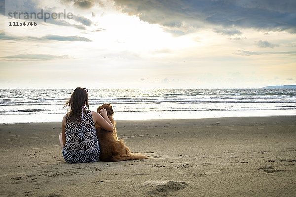 Mexiko  Nayarit  Rückansicht einer jungen Frau  die neben ihrem Hund am Strand sitzt.