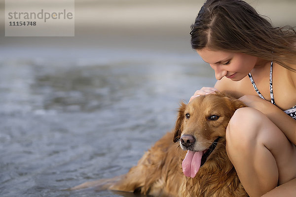 Nahaufnahme einer jungen Frau im Bikini  die ihren Golden Retriever Hund am Strand streichelt.