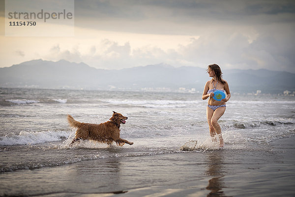 Mexiko  Nayarit  Junge Frau im Bikini beim Frisbee spielen mit ihrem Golden Retriever Hund am Strand