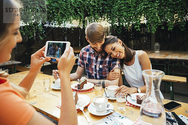 Frau macht Handyfoto von einem Paar im Freien mit Kaffee und Kuchen