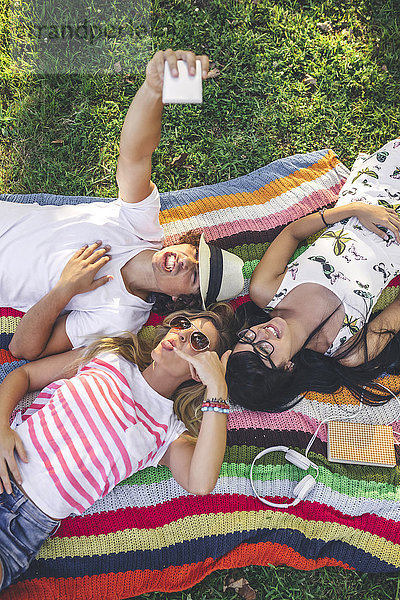 Verspielte Freunde  die auf einer Decke im Park liegen und einen Selfie nehmen.