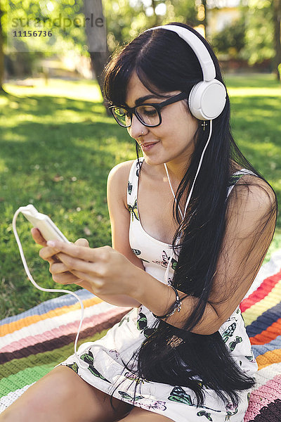 Junge Frau sitzt auf einer Decke im Park und hört Musik.