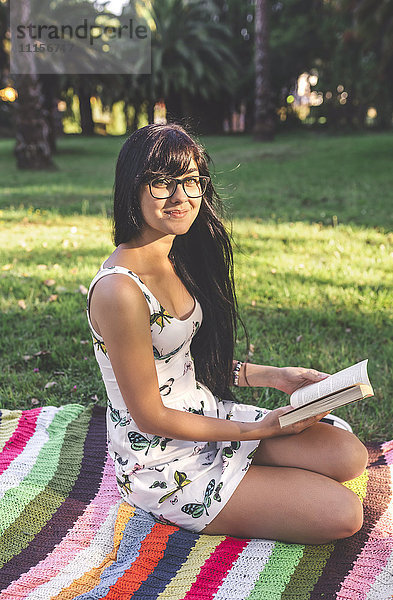 Junge Frau sitzt auf einer Decke im Park und hält ein Buch.
