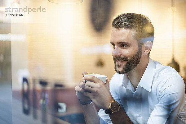 Lächelnder Geschäftsmann hält eine Tasse Kaffee hinter einer Fensterscheibe in einem Café.