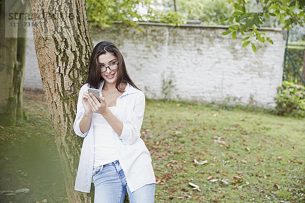 Junge Frau mit Smartphone an Baumstamm gelehnt