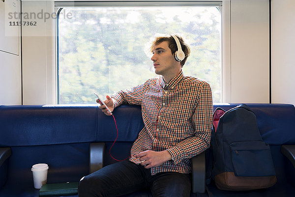 Junger Mann sitzt in einem Zug und hört Musik mit Kopfhörern.