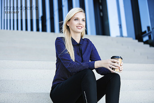 Lächelnde blonde Geschäftsfrau sitzt auf der Treppe mit Kaffee zum Mitnehmen