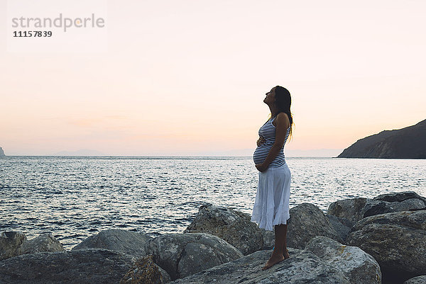 Schwangere Frau  die bei Sonnenuntergang auf einem Felsen vor dem Meer steht.