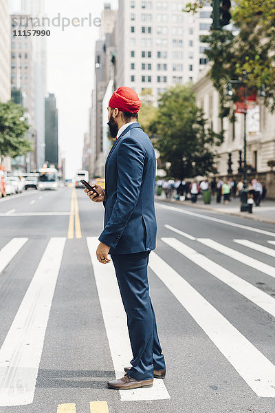 Indischer Geschäftsmann in Manhattan steht auf Zebrastreifen und hält ein Smartphone.