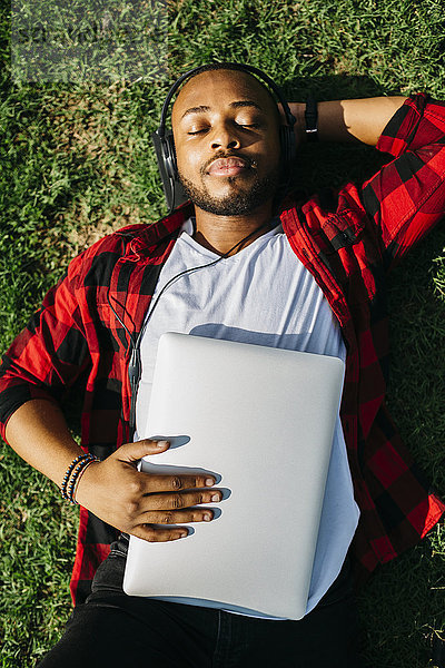 Mann im Gras liegend mit Laptop und Musik hören