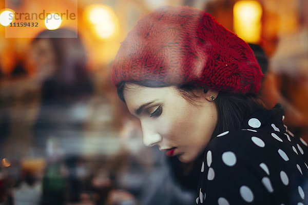 Porträt einer jungen Frau mit rotem Hut hinter der Fensterscheibe einer Kneipe am Abend