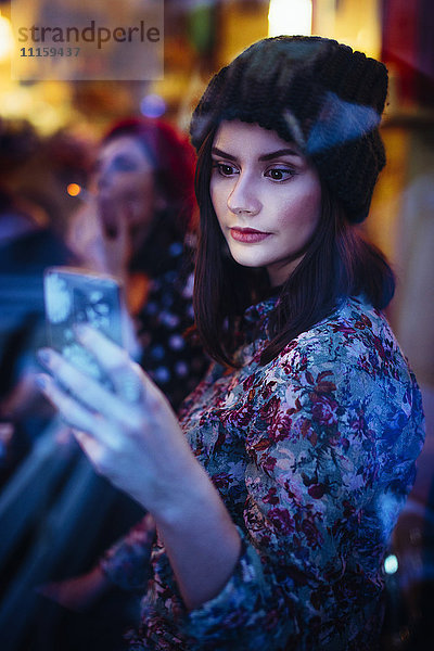 Porträt einer jungen Frau mit Smartphone hinter der Fensterscheibe einer Kneipe am Abend