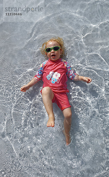Bekleidetes kleines Mädchen mit Sonnenbrille auf dem Rücken im Wasser liegend