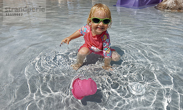 Bekleidetes kleines Mädchen mit Sonnenbrille  das im Wasser spielt.