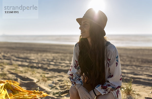 Lächelnde junge Frau sitzt am Strand bei Gegenlicht