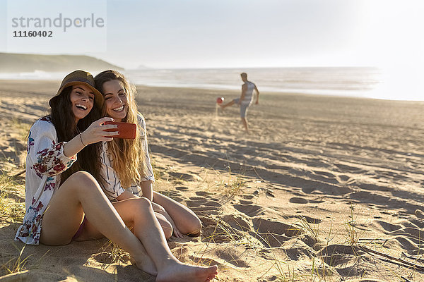 Zwei beste Freunde sitzen am Strand und nehmen Selfie mit dem Smartphone.