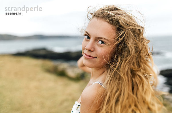 Porträt eines lächelnden blonden Mädchens mit blasenden Haaren an der Küste