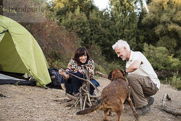 Seniorenpaar mit Hund am Zelt bei der Vorbereitung eines Lagerfeuers