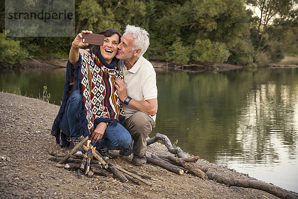 Fröhliches Seniorenpaar am See mit einem Selfie am Lagerfeuer