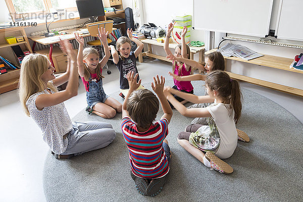Lehrer und Schüler sitzen im Kreis auf dem Boden ihres Klassenzimmers und heben die Hände.
