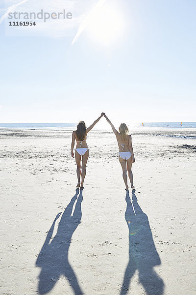 Zwei Freundinnen im Bikini gehen Hand in Hand am Strand.