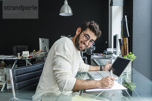 Porträt eines lächelnden jungen Mannes  der mit einem Laptop am Schreibtisch in einem modernen Büro arbeitet.