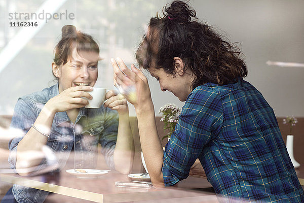 Zwei lachende Frauen sitzen hinter der Fensterscheibe eines Cafés.