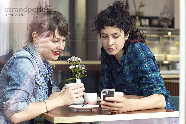 Zwei Frauen sitzen hinter der Fensterscheibe eines Coffeeshops und schauen auf das Smartphone.