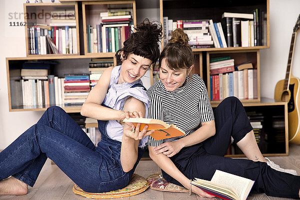 Zwei beste Freunde sitzen Rücken an Rücken auf dem Boden des Wohnzimmers und schauen sich ein Buch an.