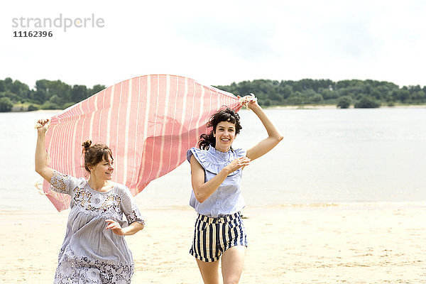 Zwei glückliche Freunde laufen Seite an Seite am Strand und halten das Tuch.