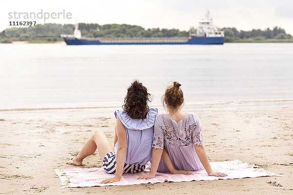 Rückansicht von zwei Freunden  die nebeneinander am Strand sitzen und ein Schiff beobachten.