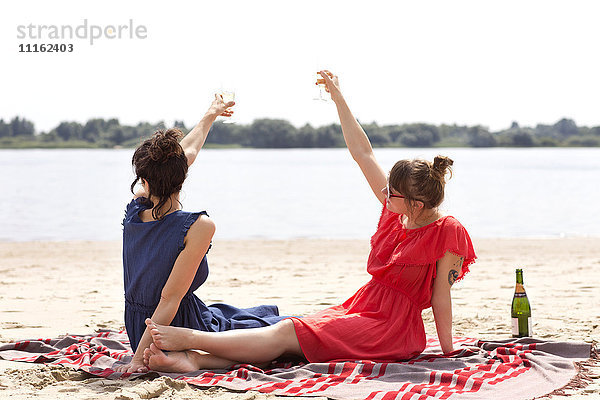 Zwei Freunde sitzen am Strand und stoßen mit einem Glas Sekt an.