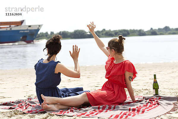 Zwei Freunde sitzen am Strand und stoßen mit einem Glas Sekt an.