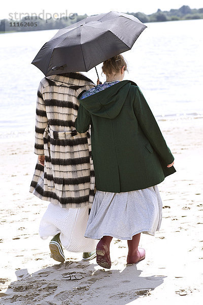 Rückansicht von zwei Freunden  die Seite an Seite mit einem Regenschirm am Strand spazieren gehen.