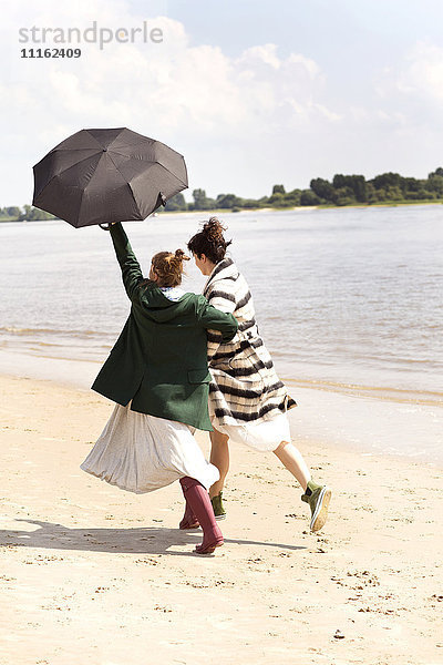 Rückansicht von zwei Freunden  die Seite an Seite am Strand mit einem Regenschirm laufen.