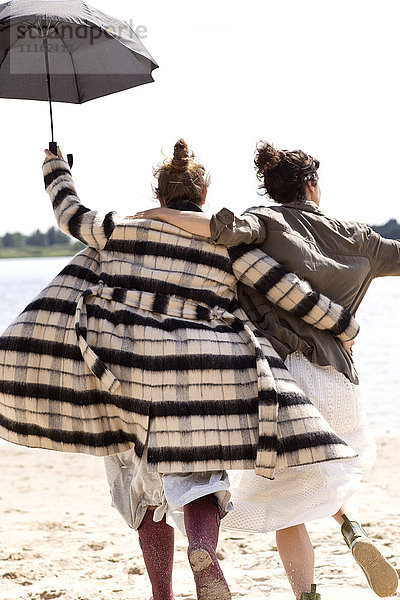 Rückansicht von zwei Freunden  die Seite an Seite am Strand mit einem Regenschirm laufen.