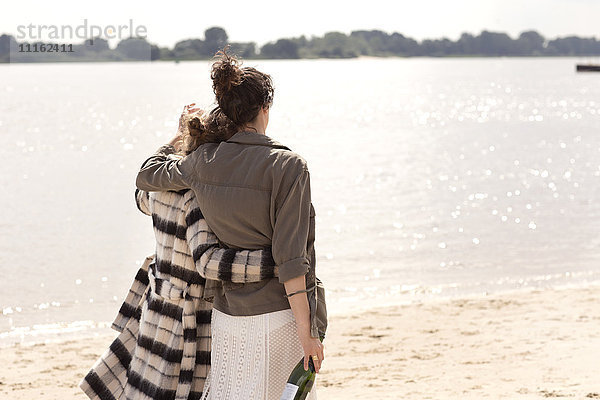 Rückansicht von zwei Freunden  die Arm in Arm am Strand mit einer Flasche Sekt spazieren gehen.