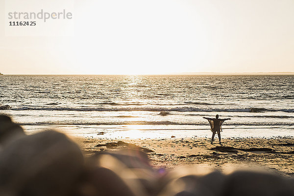 Frankreich  Halbinsel Crozon  Frau steht bei Sonnenuntergang am Strand  Arme ausgestreckt