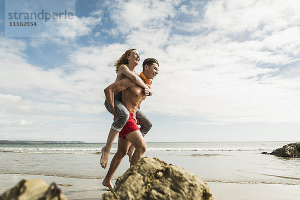 Fröhlicher junger Mann trägt Freundin Huckepack am Strand