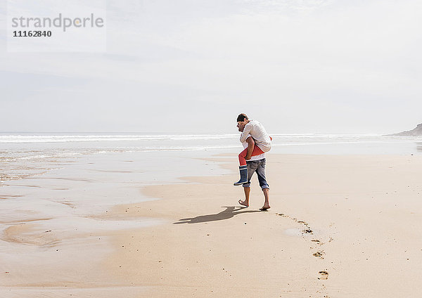 Erwachsener Mann  der seine Frau huckepack am Strand trägt.