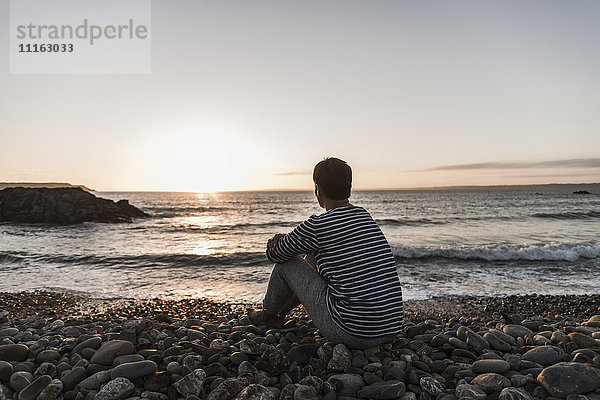 Frankreich  Bretagne  Halbinsel Crozon  Frau am steinigen Strand bei Sonnenuntergang sitzend