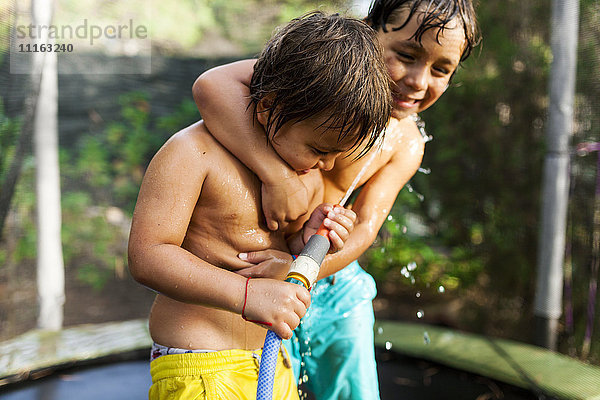 Zwei kleine Brüder spielen mit einem Wasserschlauch  der auf einem Trampolin steht.