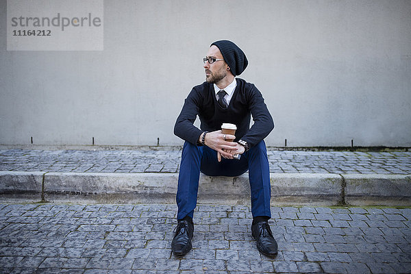 Junger Mann auf dem Bordstein sitzend mit Kaffee zum Mitnehmen