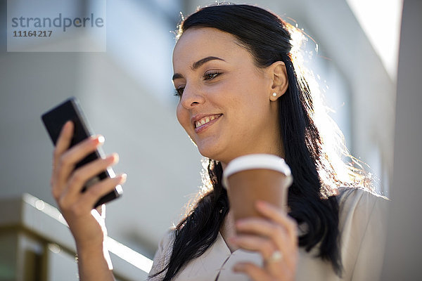 Lächelnde Frau mit Kaffee zum Mitnehmen beim Blick aufs Handy