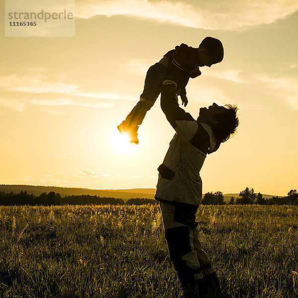 Mari Mann hebt Sohn im Feld bei Sonnenuntergang