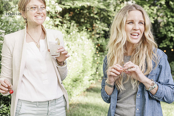 Zwei lächelnde blonde Frauen stehen in einem Garten und halten ein Getränk in der Hand.
