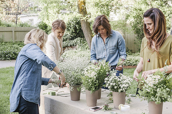 Vier Frauen  die an einem Tisch in einem Garten stehen und Blumenkränze machen.