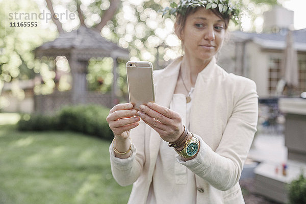Frau mit einem Blumenkranz im Haar  die in einem Garten steht und einen Selfie mit ihrem Mobiltelefon mitnimmt.