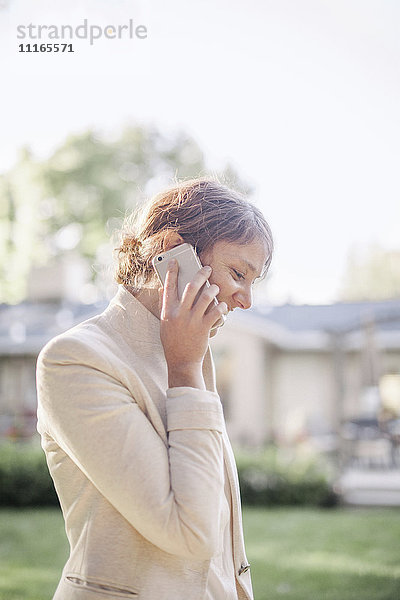Frau  die in einem sonnenbeschienenen Garten steht und mit ihrem Handy telefoniert.