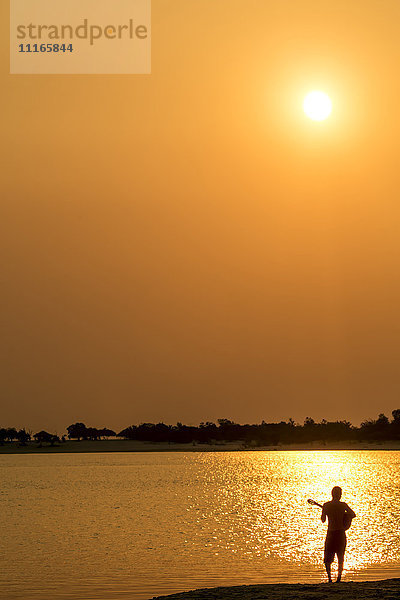 Silhouette eines kaukasischen Mannes  der am Strand bei Sonnenuntergang Gitarre spielt